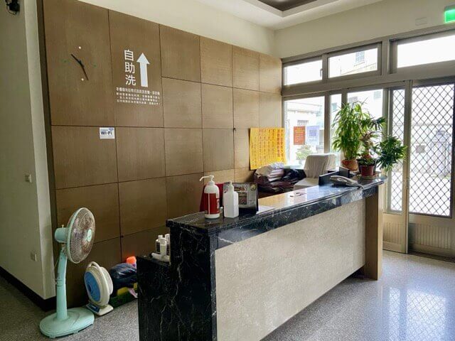 櫃檯上有綠色植栽和住宿價目表，櫃檯內有電扇和時鐘