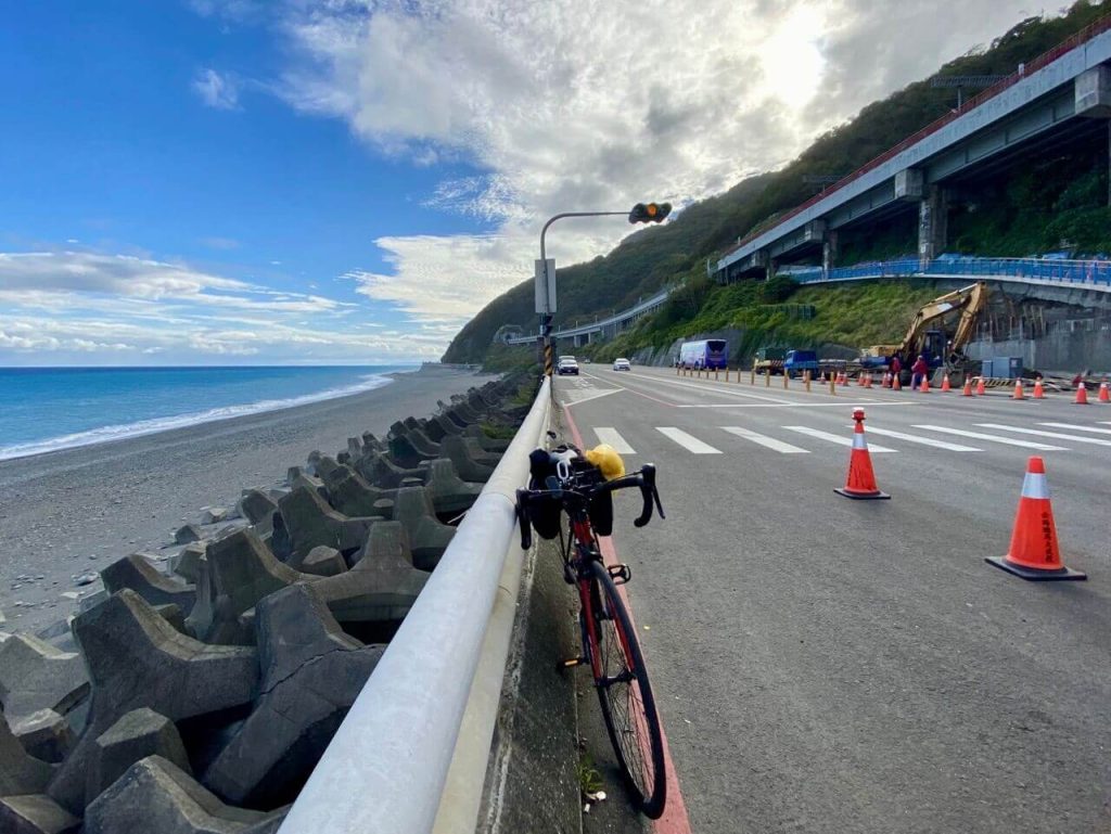 左側是大海和消波塊，右側是公路、高架鐵路和山，一台單車停靠在沿海公路的圍欄邊